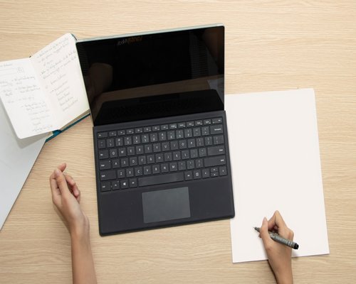 A person taking notes on a paper notebook next to a closed modern silver laptop